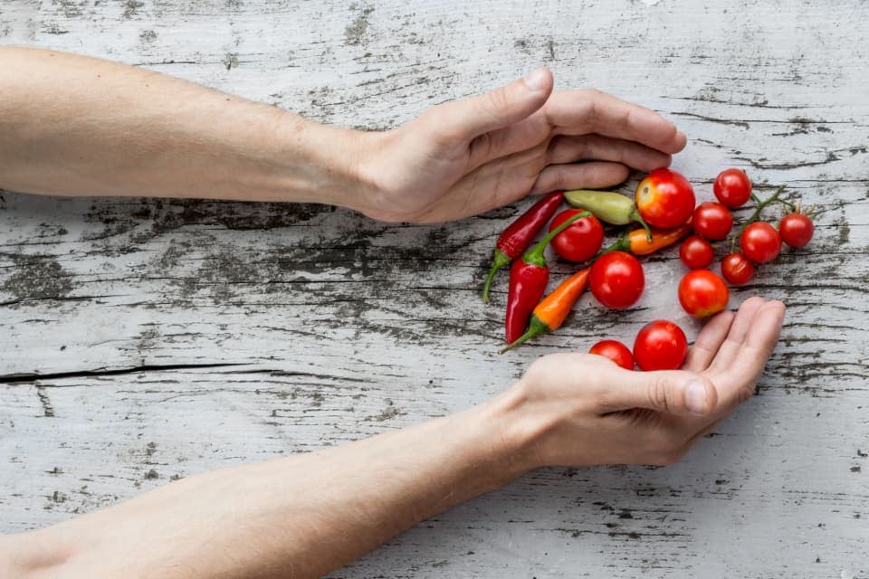 Unas manos entregan tomates y chiles frescos a otras manos, sobre una mesa de madera blanca.