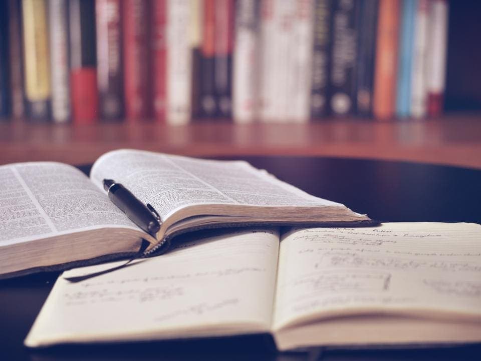 Un libro abierto y un cuaderno con apuntes a mano sobre una mesa, con una estantería de libros al fondo.