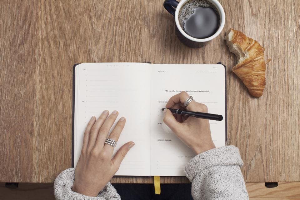 Unas manos escriben en una libreta abierta sobre una mesa de madera, junto a una taza de café y un cruasán.
