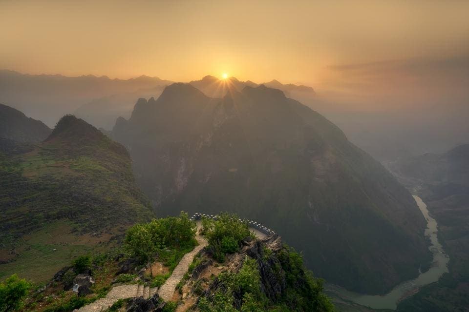 El sol asoma sobre un valle de montañas cubiertas de bruma, con un sendero que serpentea por la ladera.