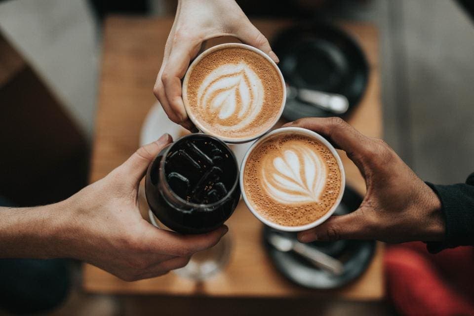 Varias manos acercan tazas de café con arte latte sobre una mesa, en un gesto de brindis.